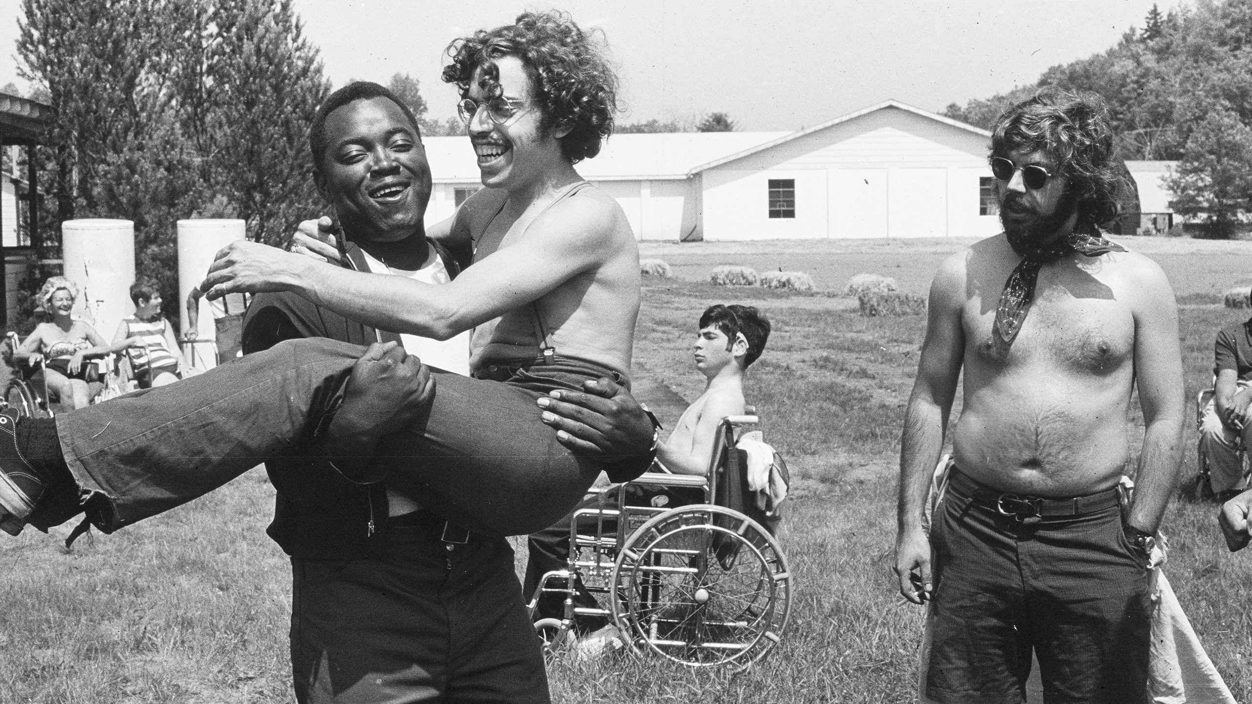 Black and white photo of a person carrying another person in the sunny outdoors. Both are laughing. A man wearing no shirt and a neck scarf looks at them. Several people in wheelchairs relax in the background.